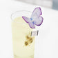 A glass cup filled with tea, featuring a butterfly-shaped tea bag hanging on the edge. The tea has a clear amber hue, with the butterfly resting delicately on the rim of the cup.