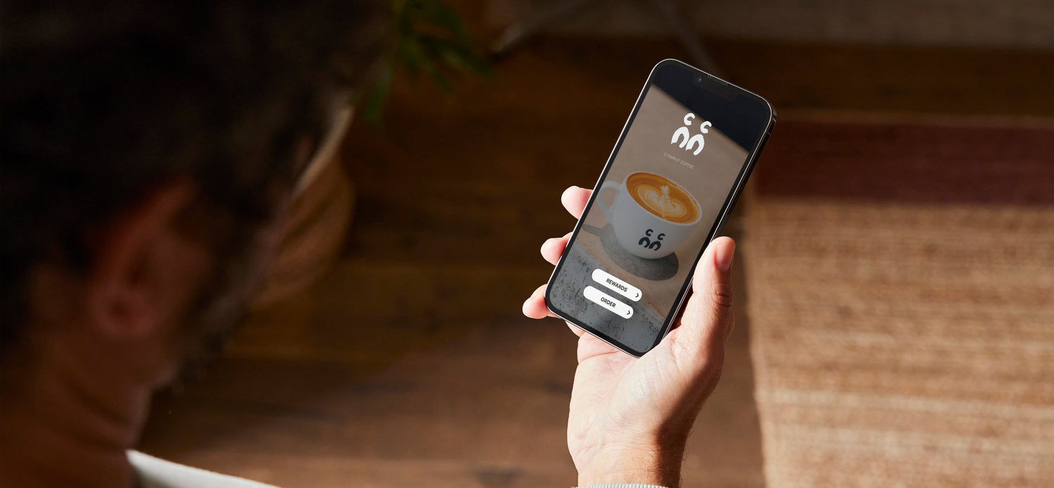 The image shows the C Market Coffee mobile app interface, where users can easily navigate to order food and beverages, earn rewards, and access exclusive offers. The clean, user-friendly design highlights key features like account management, pre-ordering, and loyalty points, making it convenient for customers to engage with the café's services and enjoy perks on-the-go.